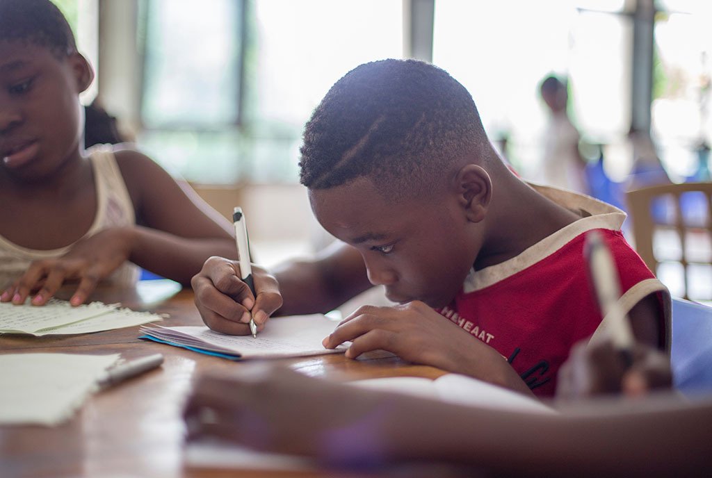 A young Black boy concentrated on writing on a paper in front of him. He is surrounded by Black classmates.