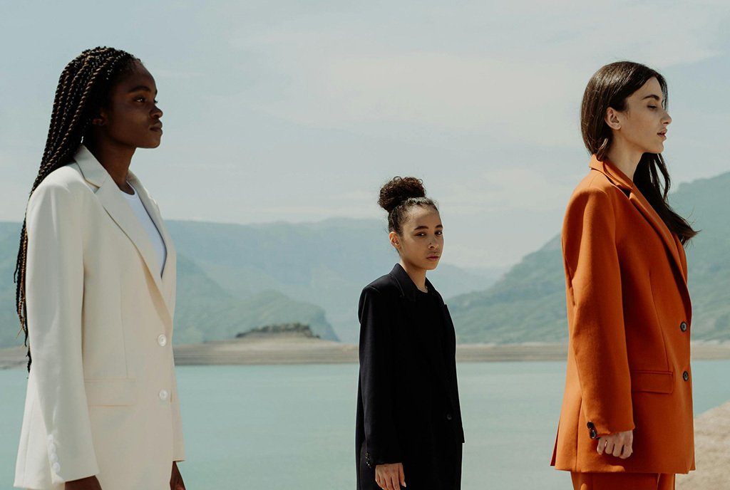 A group of three diverse woman in tailored suits facing the right and standing in front of a scene of mountains and a lake.