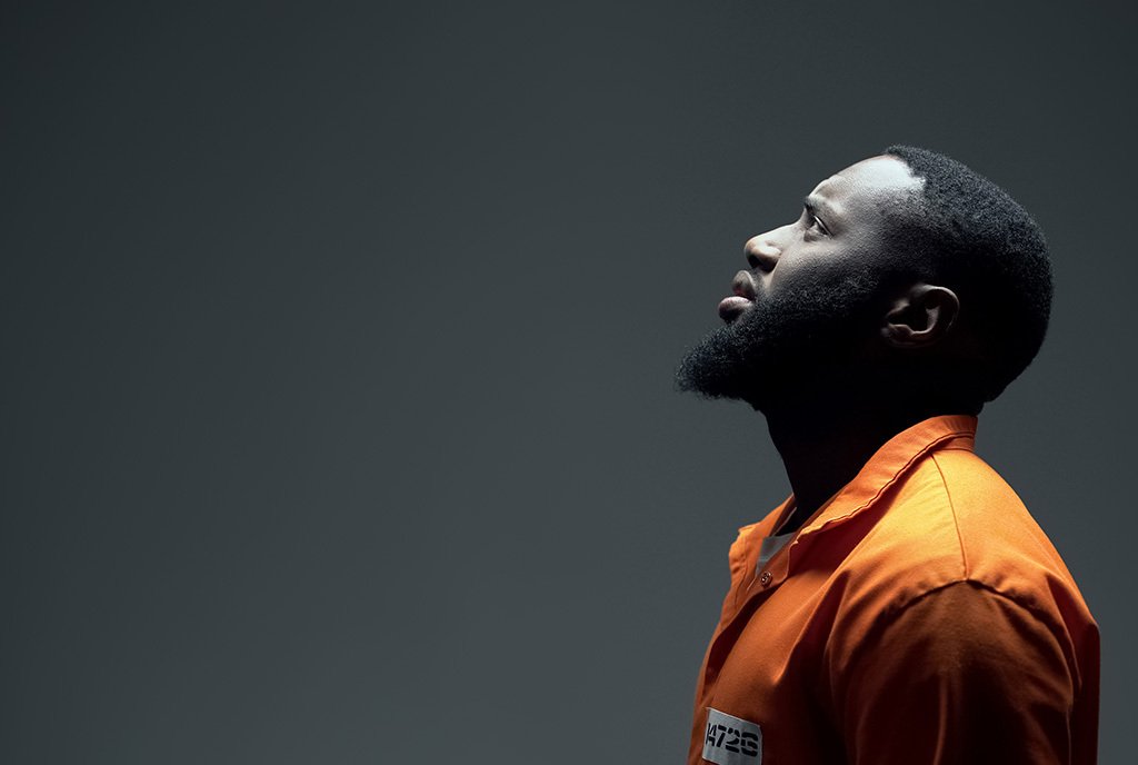 Black man in orange jumpsuit, looking up into a light.