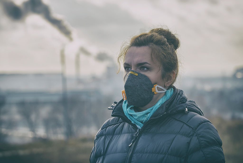  A brown-haired white woman wearing an anti-smog mask. Behind her, are smoke stacks emitting black smoke into the air.