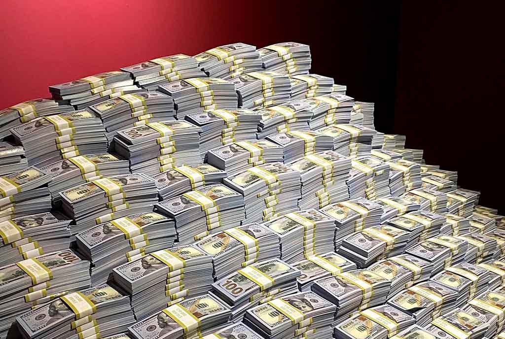An exaggerated pile of bands of money, stacked high against a red wall.
