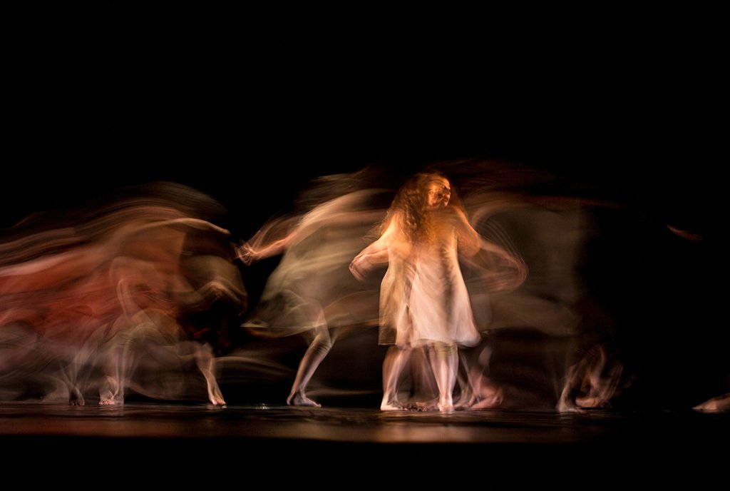 A long-exposure shot of a woman in a white dress doing an improvised dance on stage.
