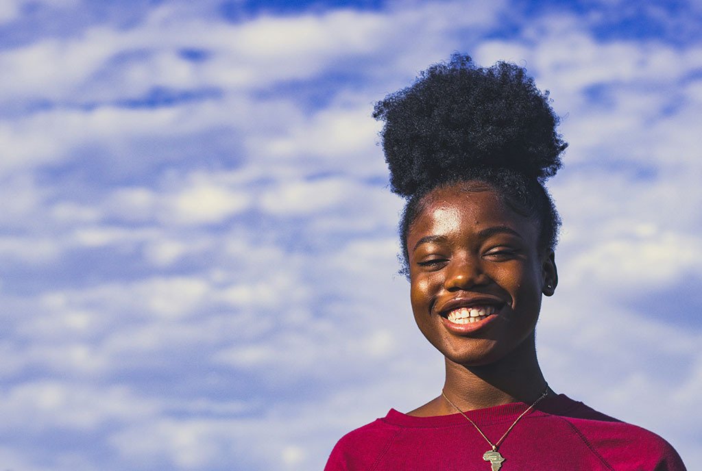 A young Black girl with an afro puff, against a blue sky, smiling widely.