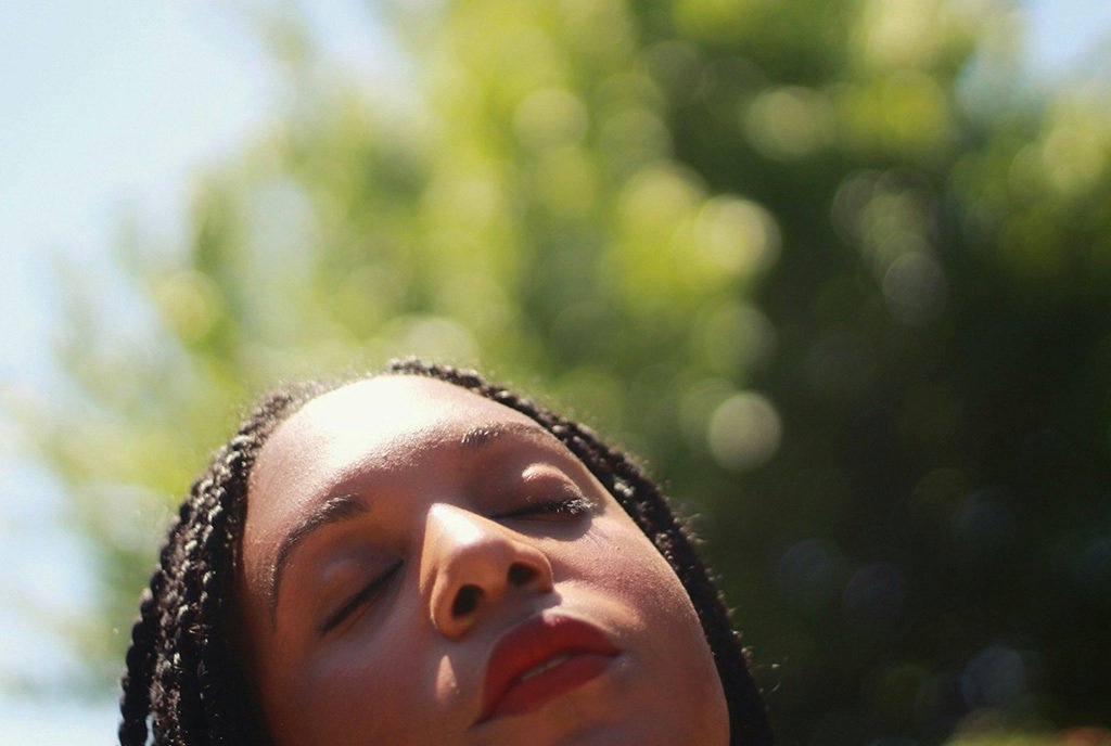 A Black woman with red lipstick and knotless braids closing her eyes and turning her face to the sun. There is a tree behind her.