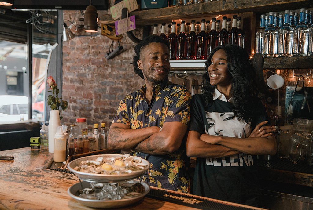 Two Black business owners, male and female, standing back to back in their bar business and smiling at each other