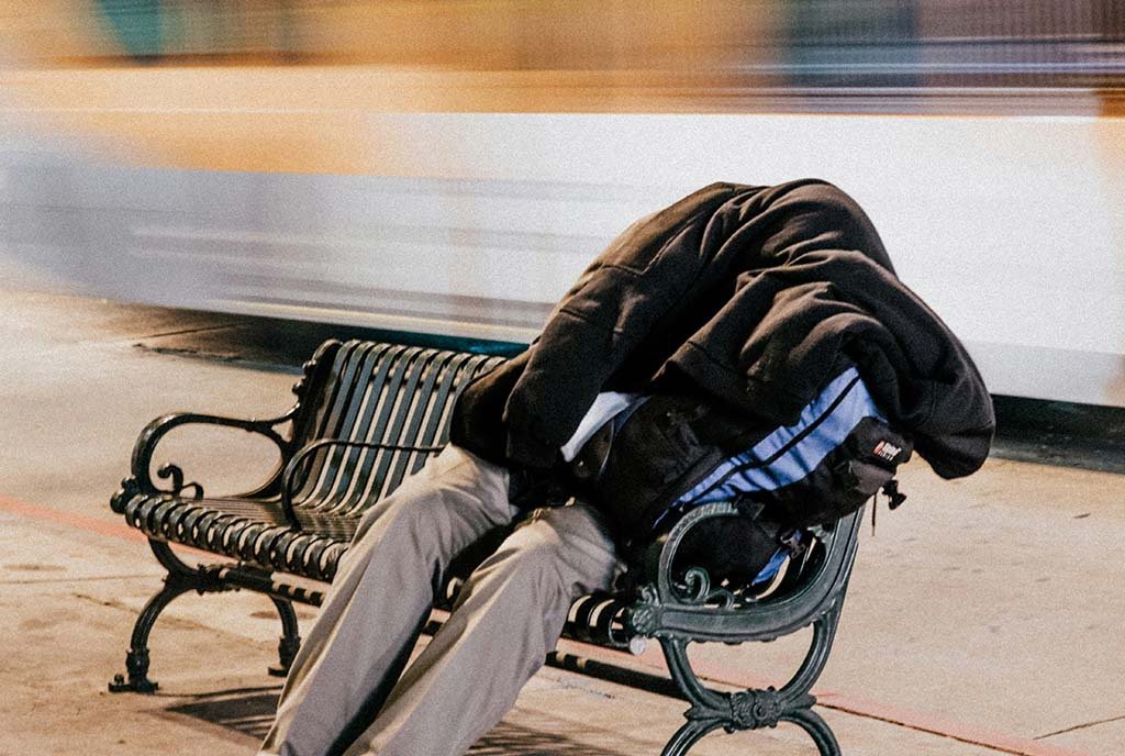 A long-exposure shot of a homeless person sleeping on a public bench, as a buss whizzes by behind them.