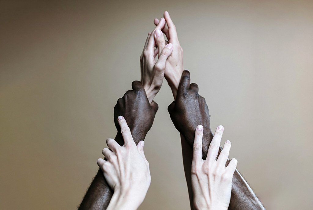 An image of a pair of white hands, holding up a pair of black hands, which are holding up another pair of white hands. The hands support each other and form a triangular shape together.