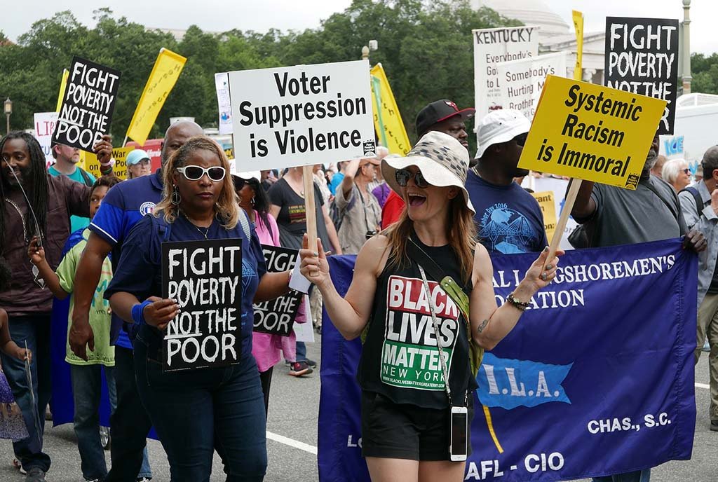 A group of protestors with the Poor People's Campaign marching down the street with signs that read “Fight Poverty, not the Poor” and “Voter Suppression is Violence”