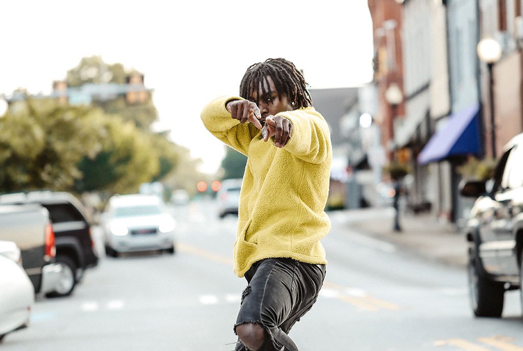 A Black man with locs dancing in the middle of an urban street, pointing at the camera and wearing a yellow sweatshirt.