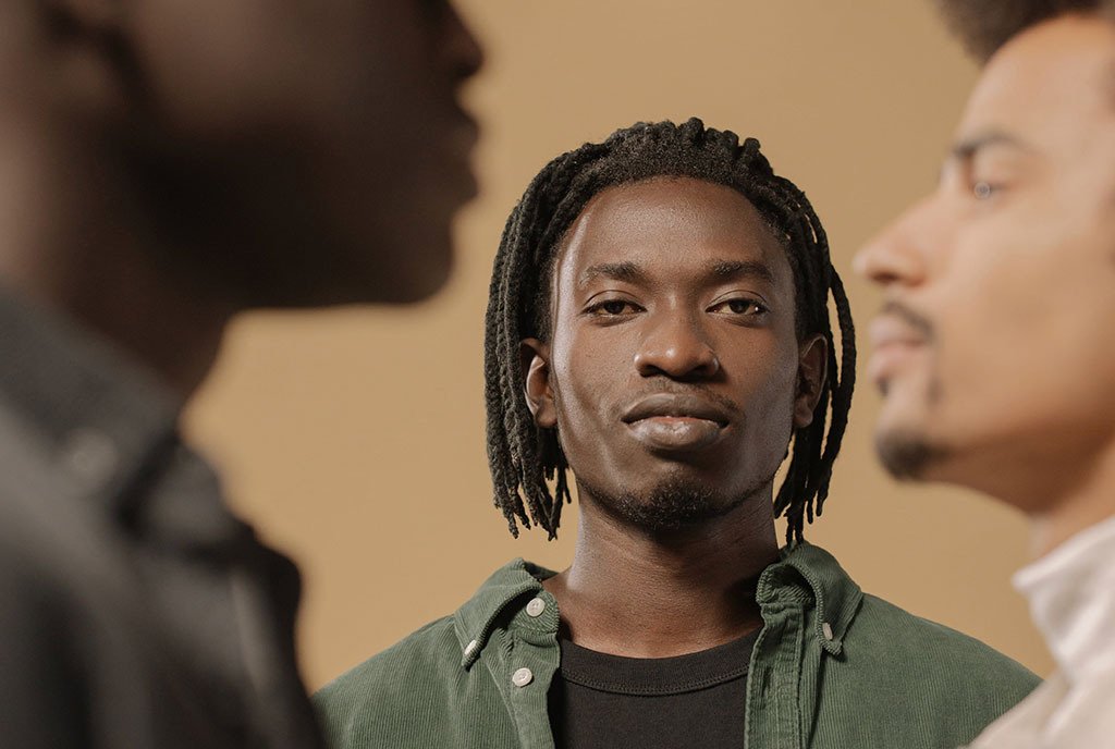 A Black man with locs looking directly into the camera with a determined look on his face. On both sides of him, there are two Black men in the foreground.