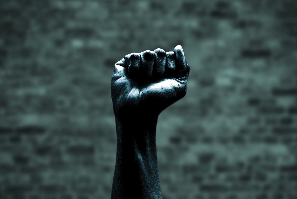 A Black fist raised in the air with a faded grey brick wall in the background. 