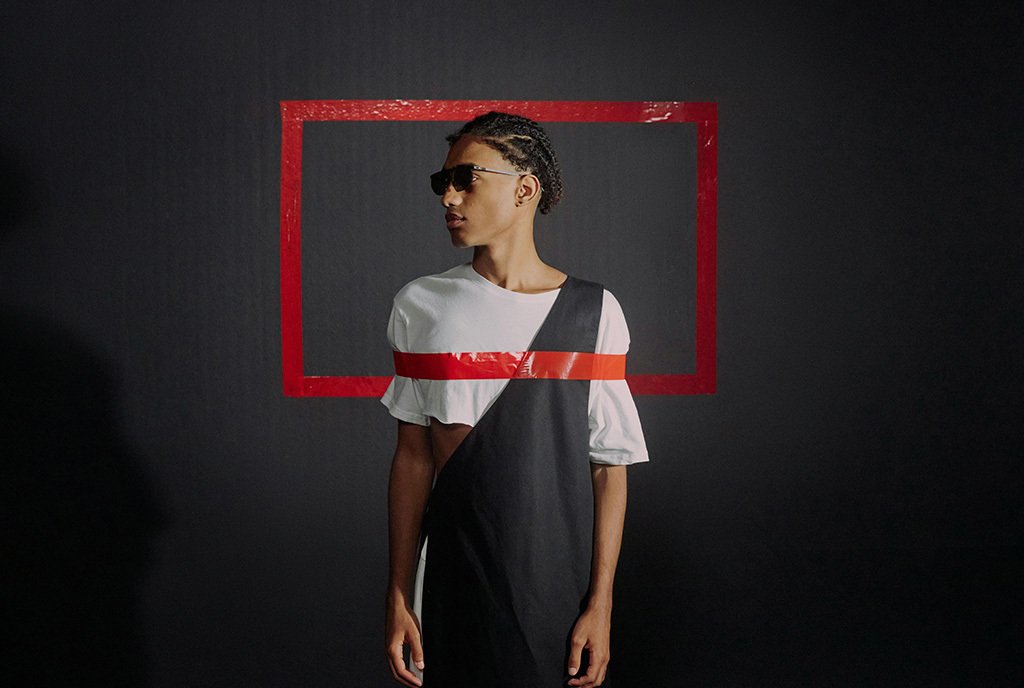 A Black man with braids and sunglasses, turning his head and showing his profile. His person is bound by a box made of red tape that crosses over his torso.