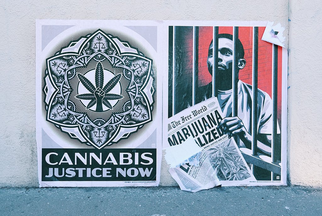 A street art work by Shepard Fairey showing a Black man behind bars reading a newspaper that says “Marijuana Legalized”. He is next to a marijuana justice design that says “Cannabis Justice Now”