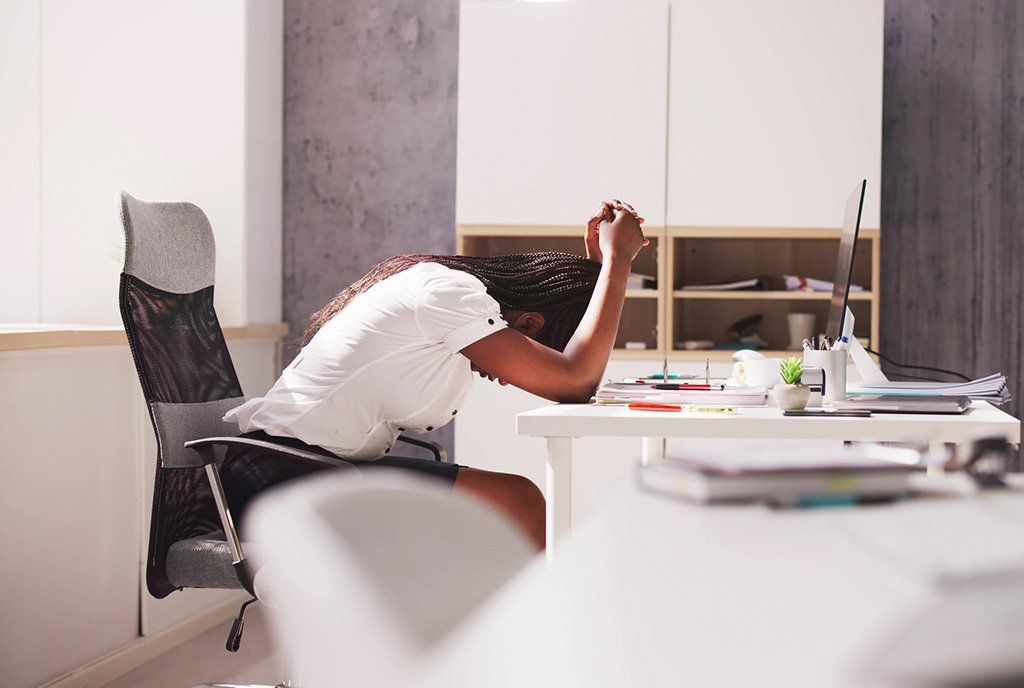 A Black woman at an office desk with her head down in frustration.