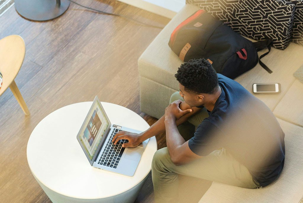 An overhead shot of a young Black man sitting on a couch, looking at his computer screen with his bookbag beside him.