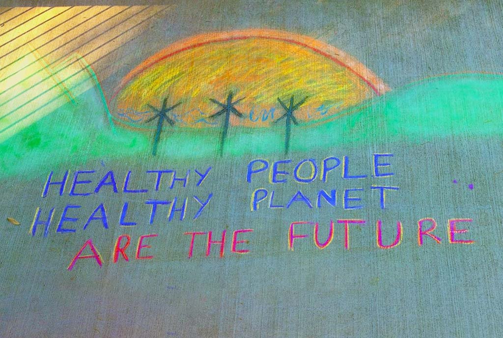 A sidewalk chalk drawing that reads, “Healthy People, Healthy Planet are the future” below a drawing of wind mills in a field with a sunset.
