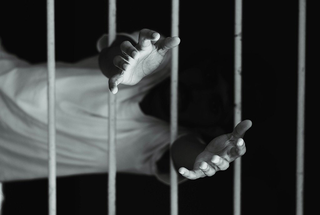 a black and white photo of a man’s hands reaching through metal bars from a dark cell.