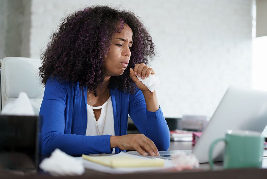A Black woman with curly hair sitting at her work station, coughing into a napkin with a miserable look on her face.
