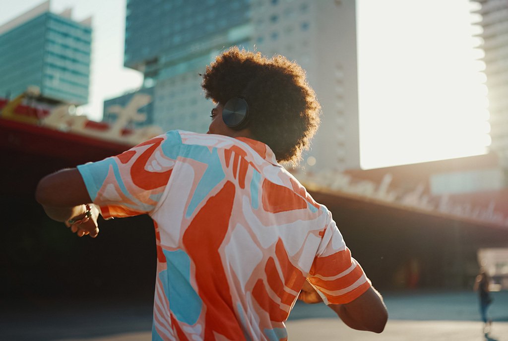 A young Black man, wearing headphones and a brightly colored shirt, dancing in a southern cityscape.