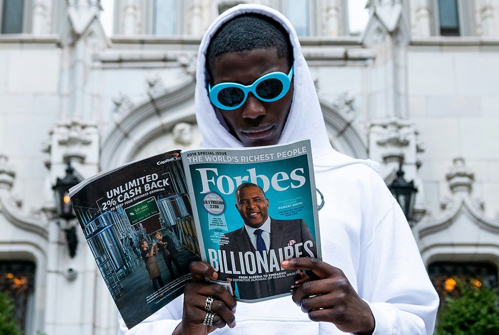 A Black man wearing sunglasses reading an issue of Forbes Magazine titled, “Billionaires: The Worlds Richest People"