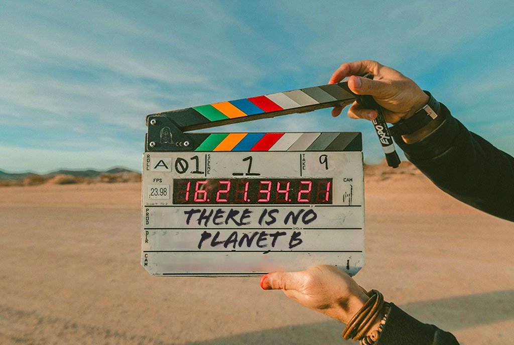 A Person holding a film clapboard in the middle of a desert landscape. The writing on the clapboard reads, “There is no planet B”