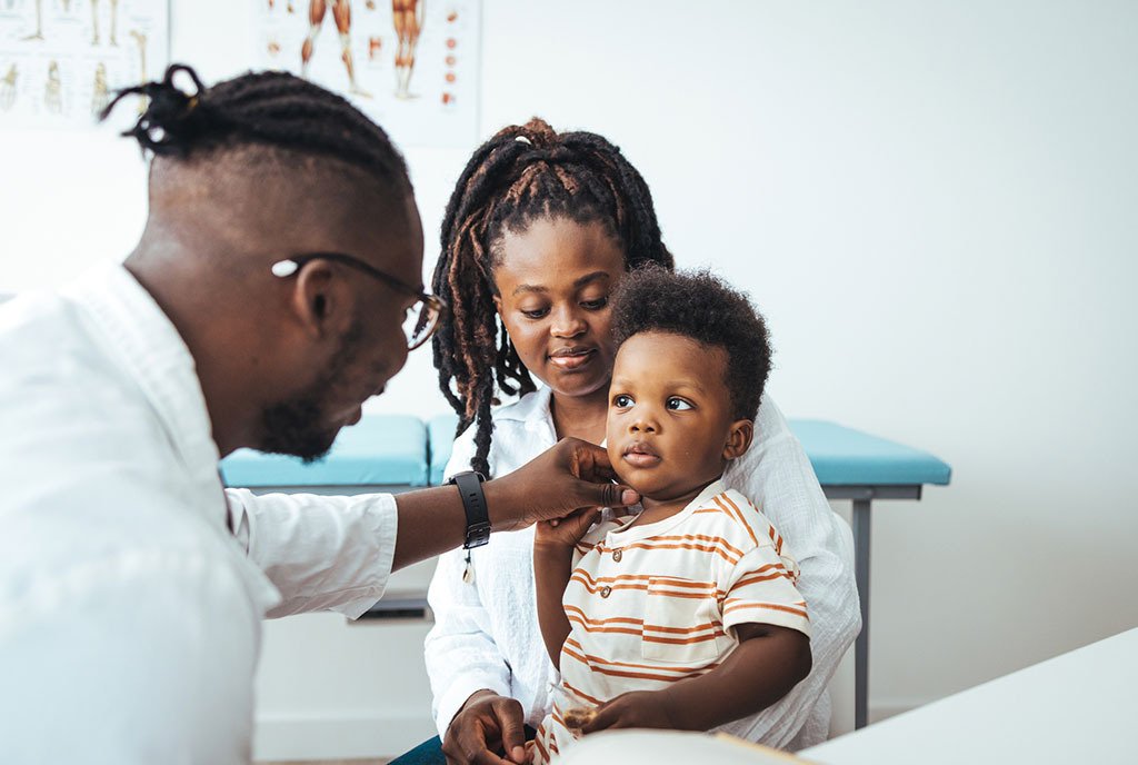 A Black pediatrician holds a stethoscope to a young Black boy’s chest, while the child sits in his mother’s lap.