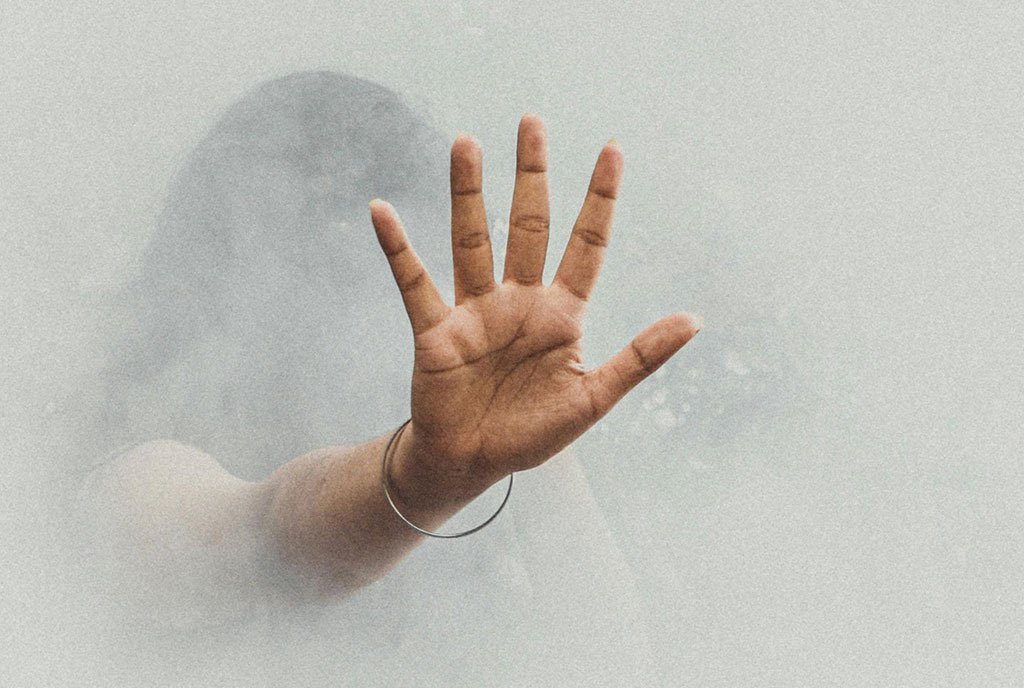A brown woman’s hand emerges from a cloud of fog. Her palm is flat and fingers are slightly spread, as if stopping something.