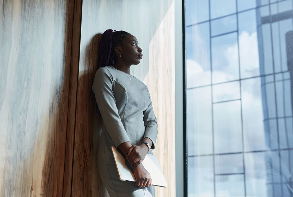 A Black woman with braids, leaning against an office wall, holding a computer and looking out the window with a disappointed look on her face