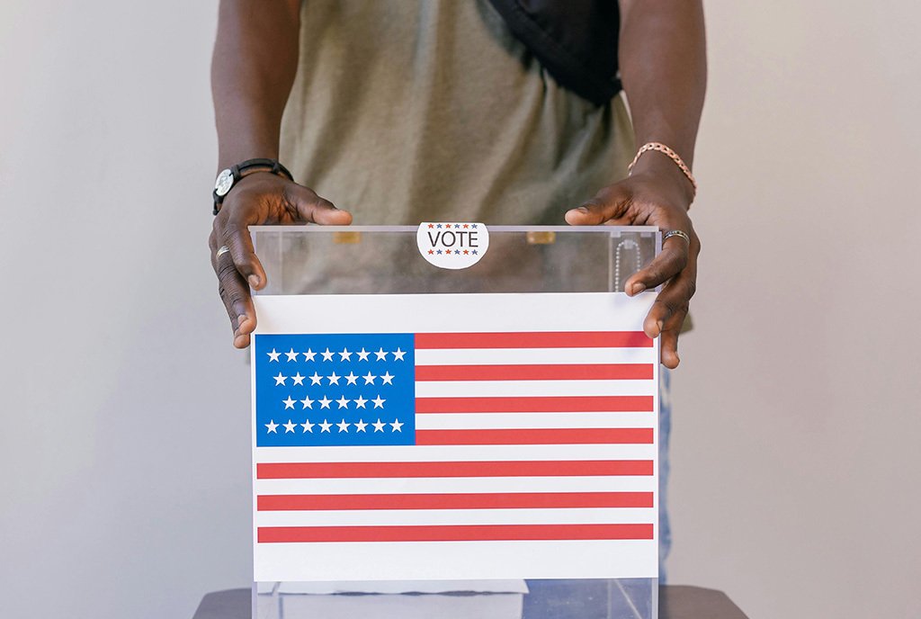 A Black man’s hands holding a transparant ballot box, with an American flag on the front.