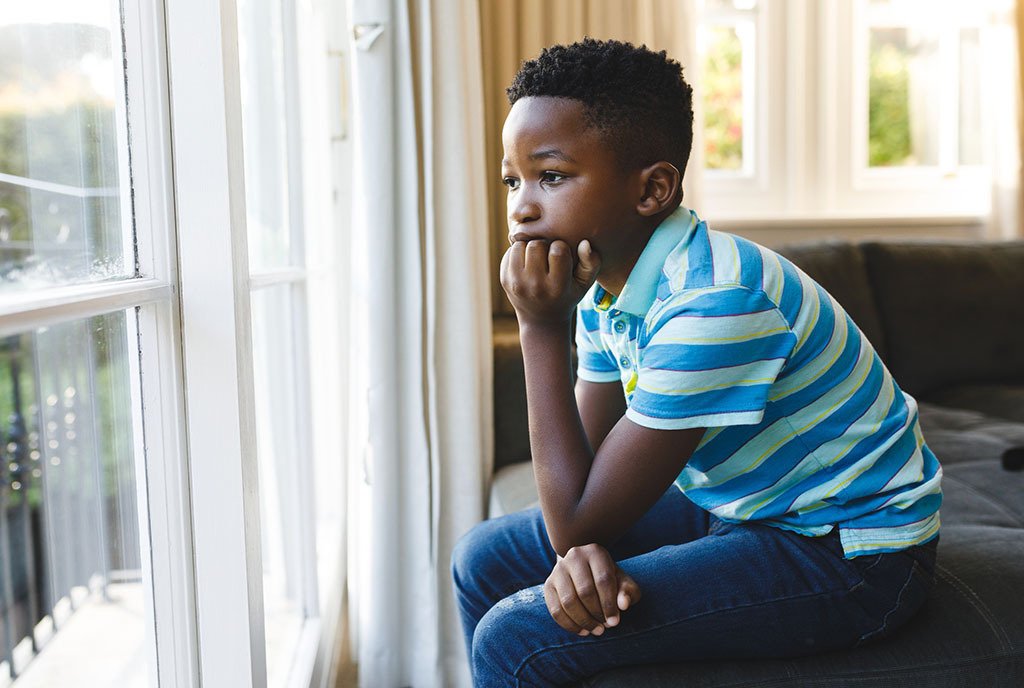 A young, Black boy sitting inside and looking out of the window with a bored expression on his face.