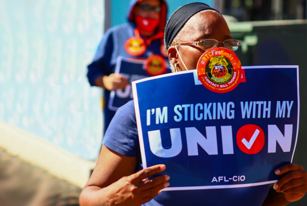 A Black woman protesting and holding a sign that reads, “I’m Sticking With My Union”