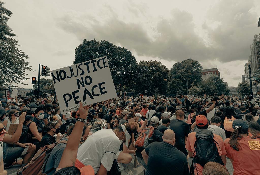 A large protesting crowd in the streets, a protestor holds up a sign that reads, “No Justice, No Peace”