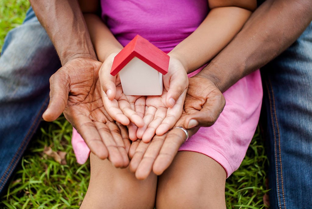A Black family sits together on the grass, holding a small wooden house in their folded hands.
