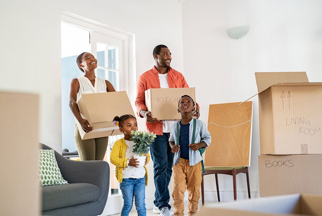 Black family with two children moving into a new house and carrying labeled cardboard boxes.