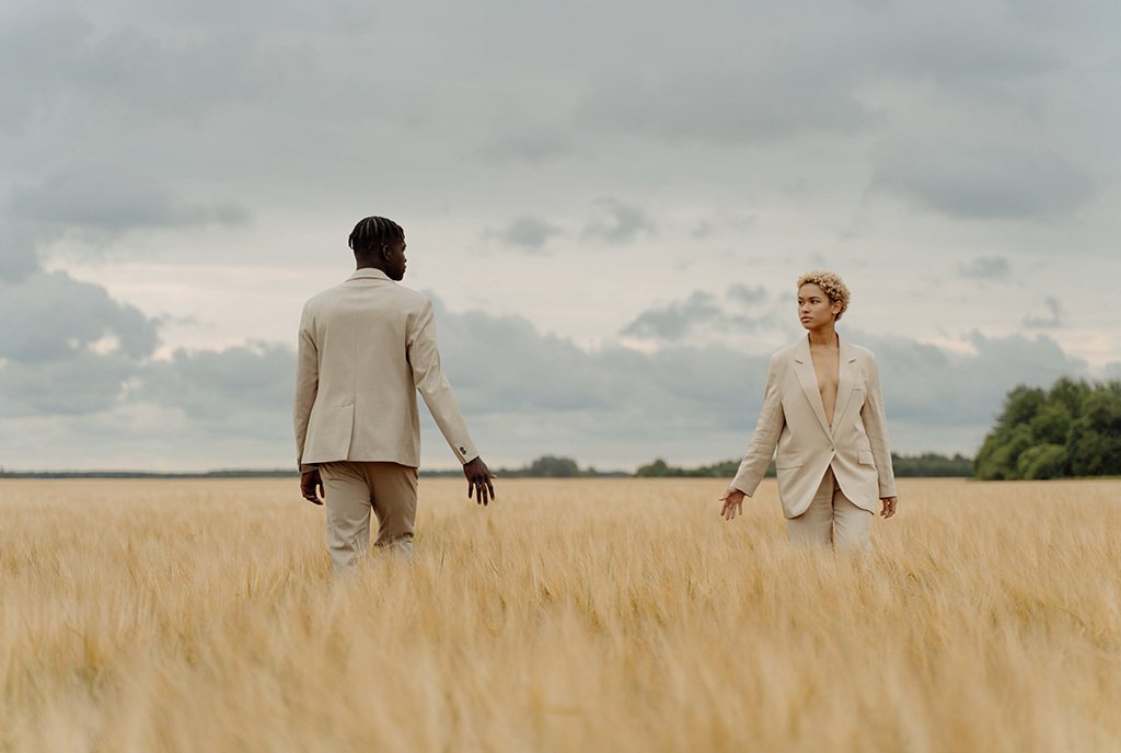 A Black man and Black woman in a field of low yellow grass, facing each other and walking by with hands extended towards each other.