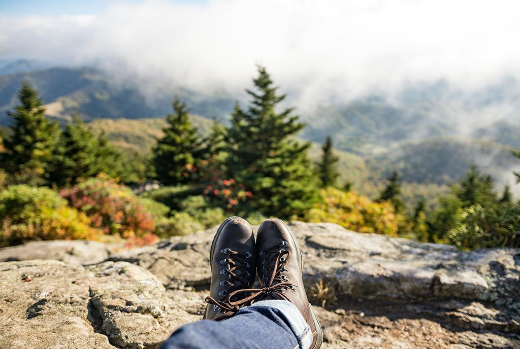 An image of a person’s feet wearing hiking boots, perched on top of a rock. There is a beautiful mountainous landscape in the background.
