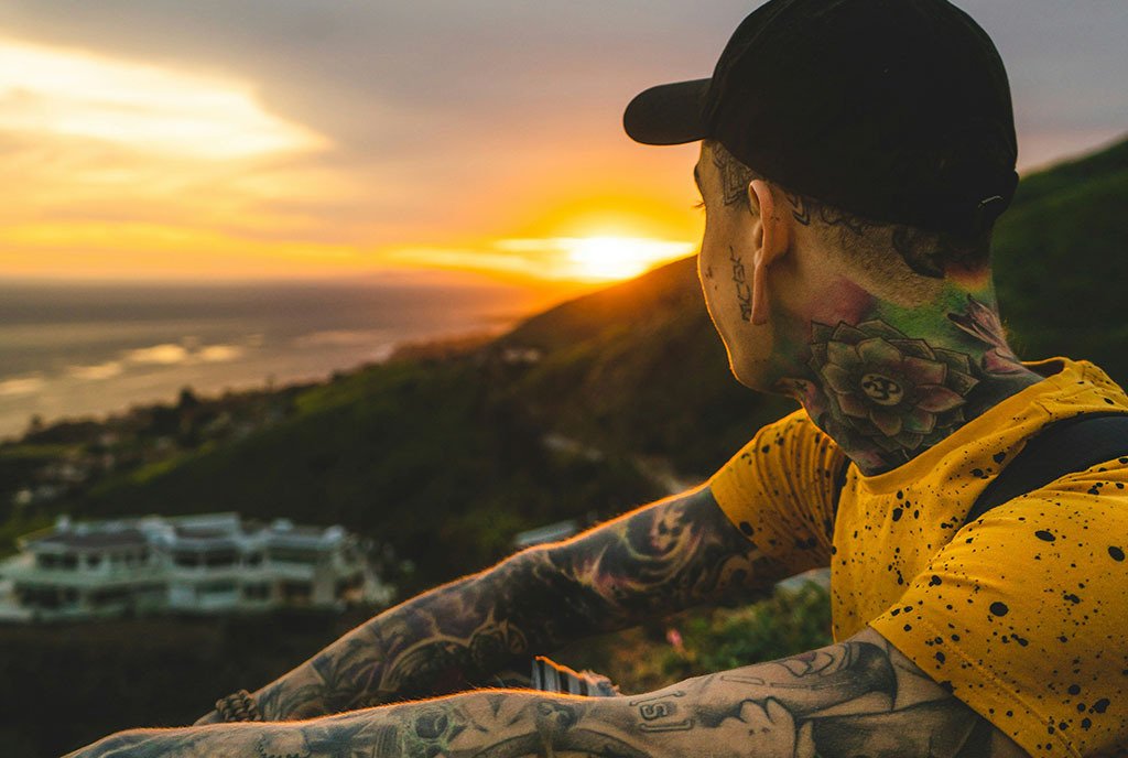 A tattooed man wearing a baseball cap, looking out over a peaceful wooded valley during sunset.