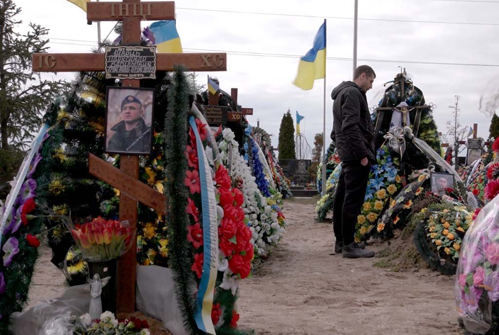 Image of a man standing in a cemetery filled with flowers. At least 11,000 Ukrainian civilians and 31,000 Ukrainian soldiers have died since the Russian invasion in February 2022. United Nations experts believe the actual death toll is “considerably higher.”