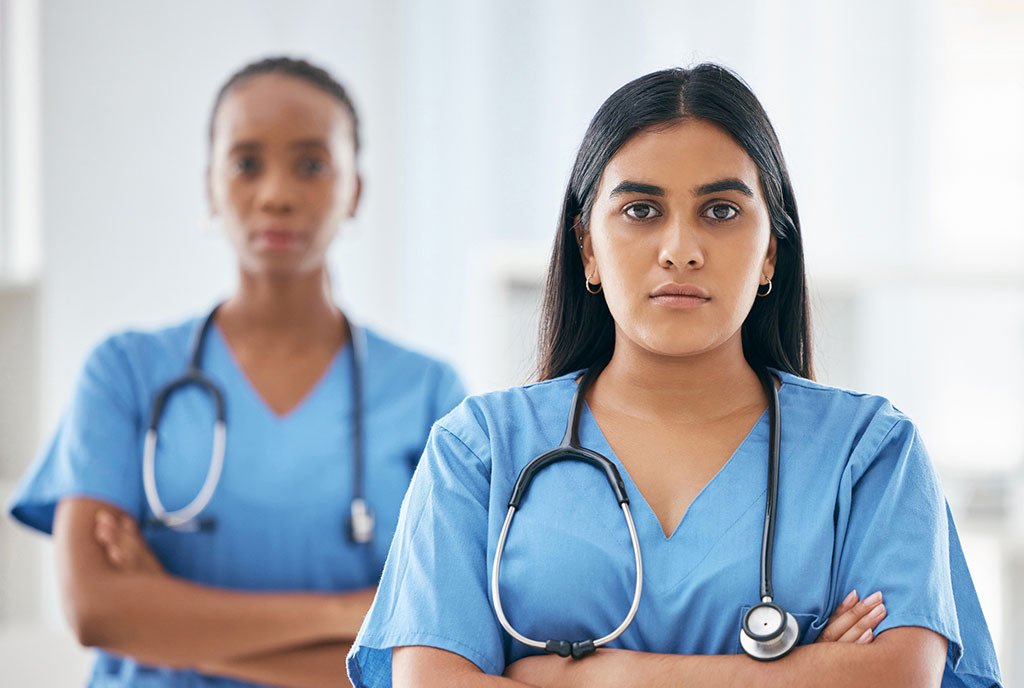 A portrait of two women medical students, one Black, and one Brown, wearing blue scrubs. They face the camera with arms crossed and serious looks on their faces.
