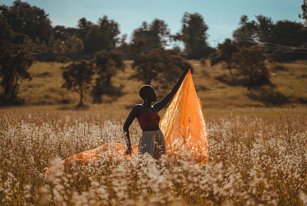 A Black woman in a flowery field, flying a bright orange sheer fabric in the wind.