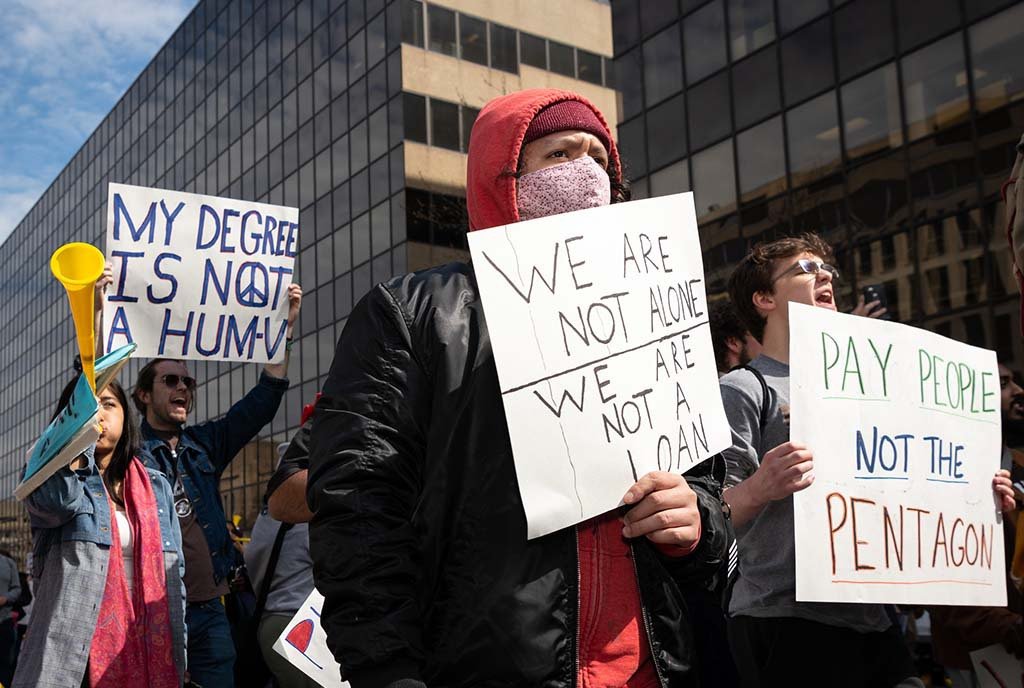 Protestors at a Student Debt protest hold up signs that read, “ We are not alone. We are not a loan” and “Pay People. Not the Pentagon” and “My Degree is not a Hum-V"