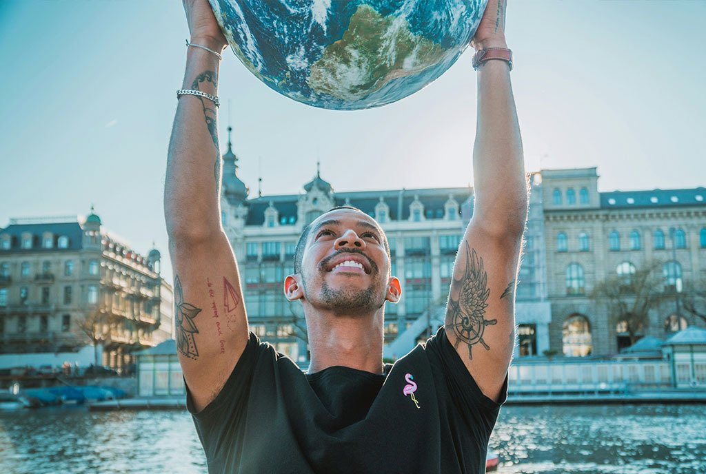 A tattooed man holds up a globe over his head with a large smile on his face.