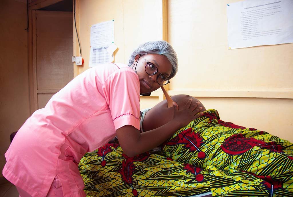 A Black midwife in pink scrubs using a wooden tool to listen to the pregnant belly of a Black woman wrapped in Ankara fabric.