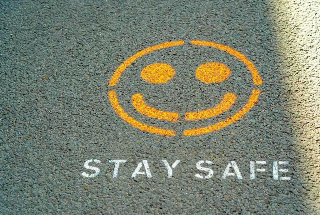 A spray painted yellow smiley face on the sidewalk, with words underneath reading, “STAY SAFE”