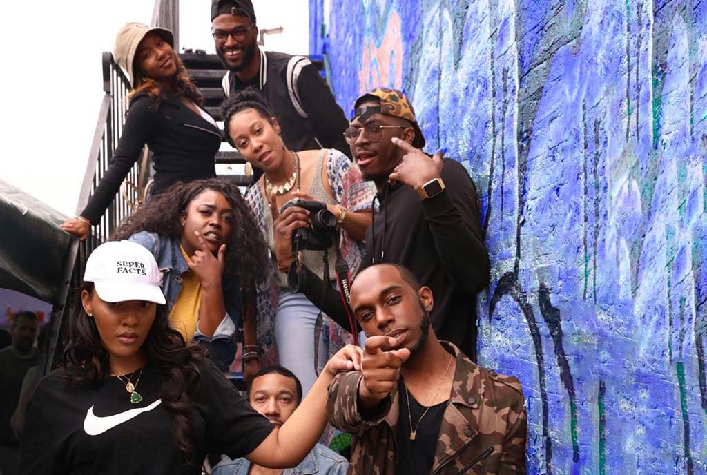 A group of Black people standing together in front of a graffiti wall and posing playfully for the camera.
