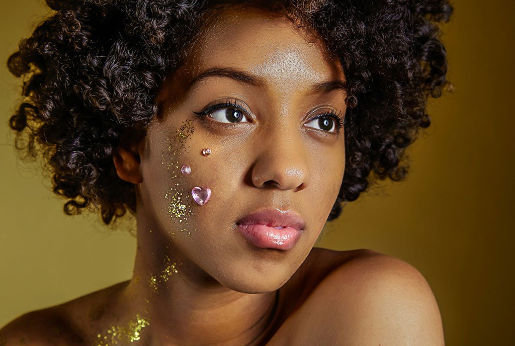 A Black woman with curly hair and glitter on the side of her face looking into the distance