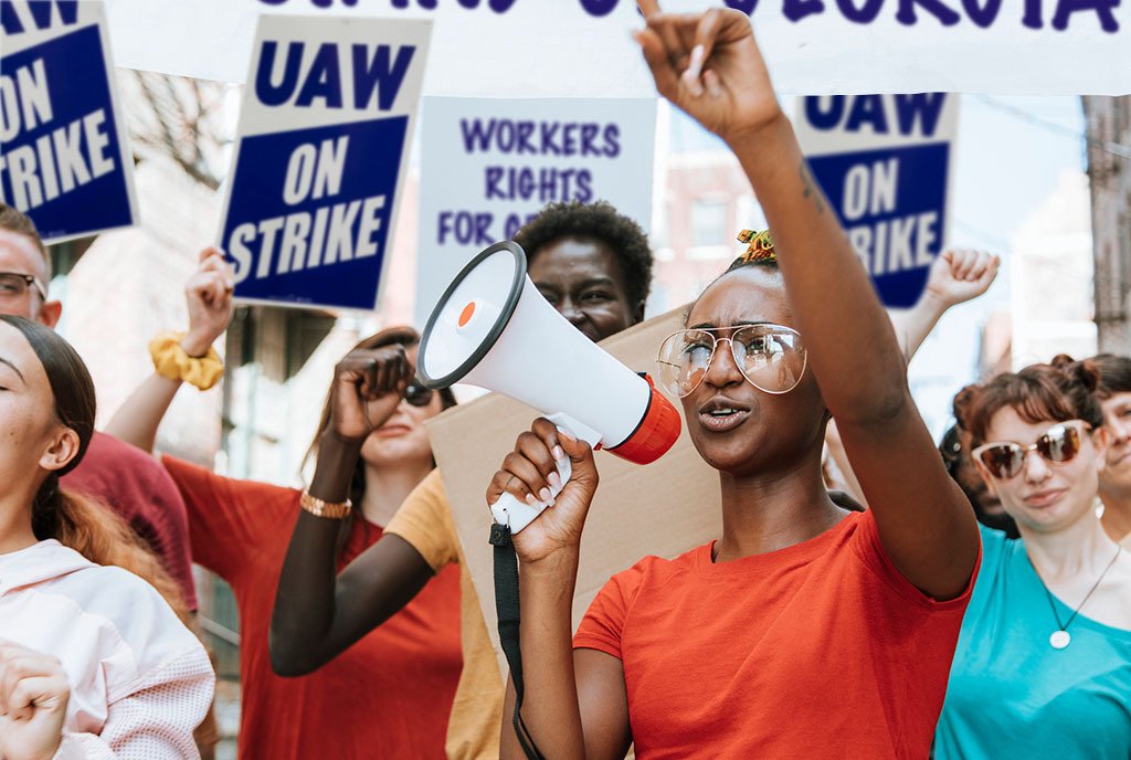 A Black women leading a United Auto Workers Protest and holding a megaphone, while a group of protestors stand behind her holding signs that read, “UAW On Strike”