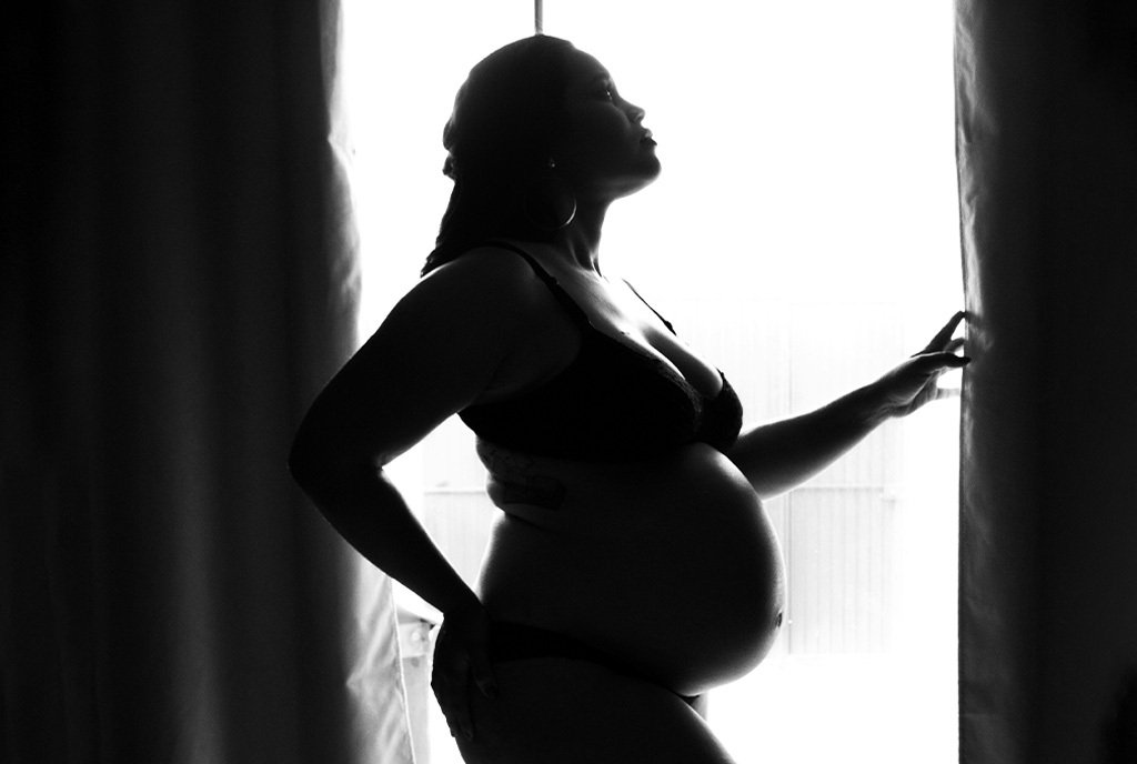 Black and white photo of a pregnant Black woman with braids standing in front of a window and looking up thoughtfully. She is wearing a black brasserie.