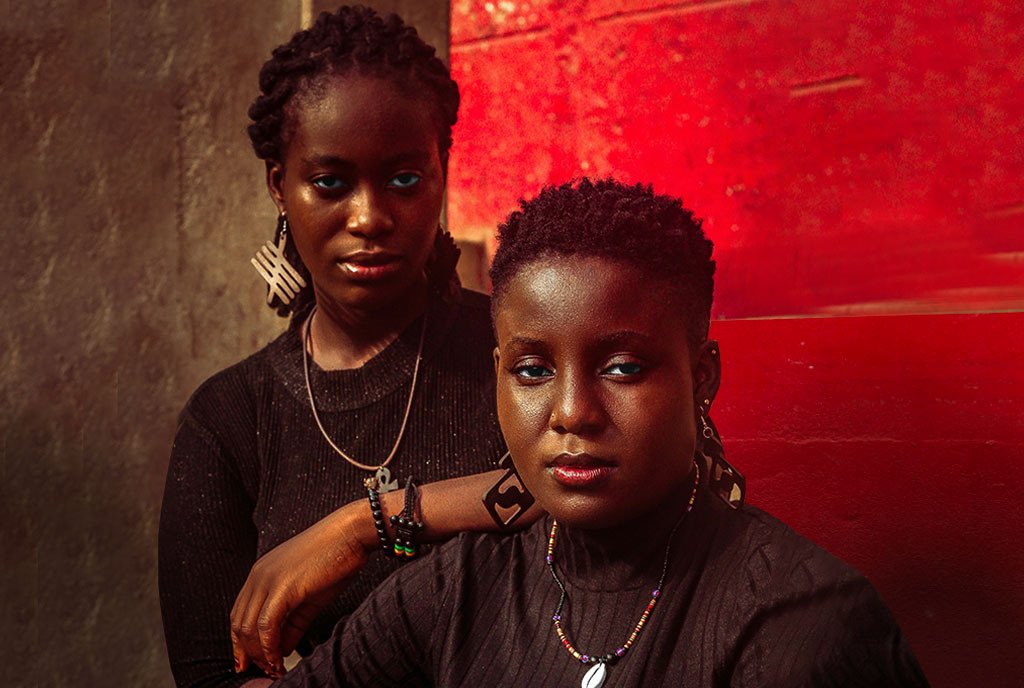 Two cool Ghanaian women wearing traditional, adinkra-themed jewelry. One woman is leaning on the other as they look into the camera with relaxed expressions.