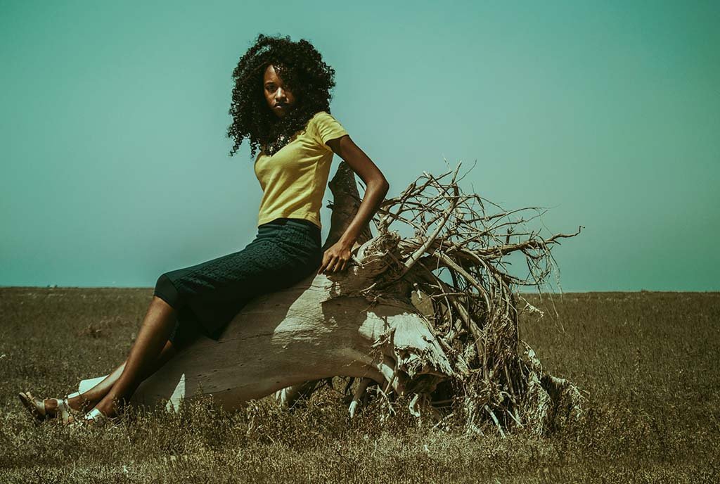 A young Black woman leaning on a fallen tree trunk and looking into the camera. The field behind her shows environmental degradation with dried grass.